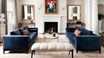 Living room with blue velvet sofas oriental rug fire lit and colorful artwork over mantel mirrors in aloves classical pillar detail and bust statue multipaned windows and white ottoman