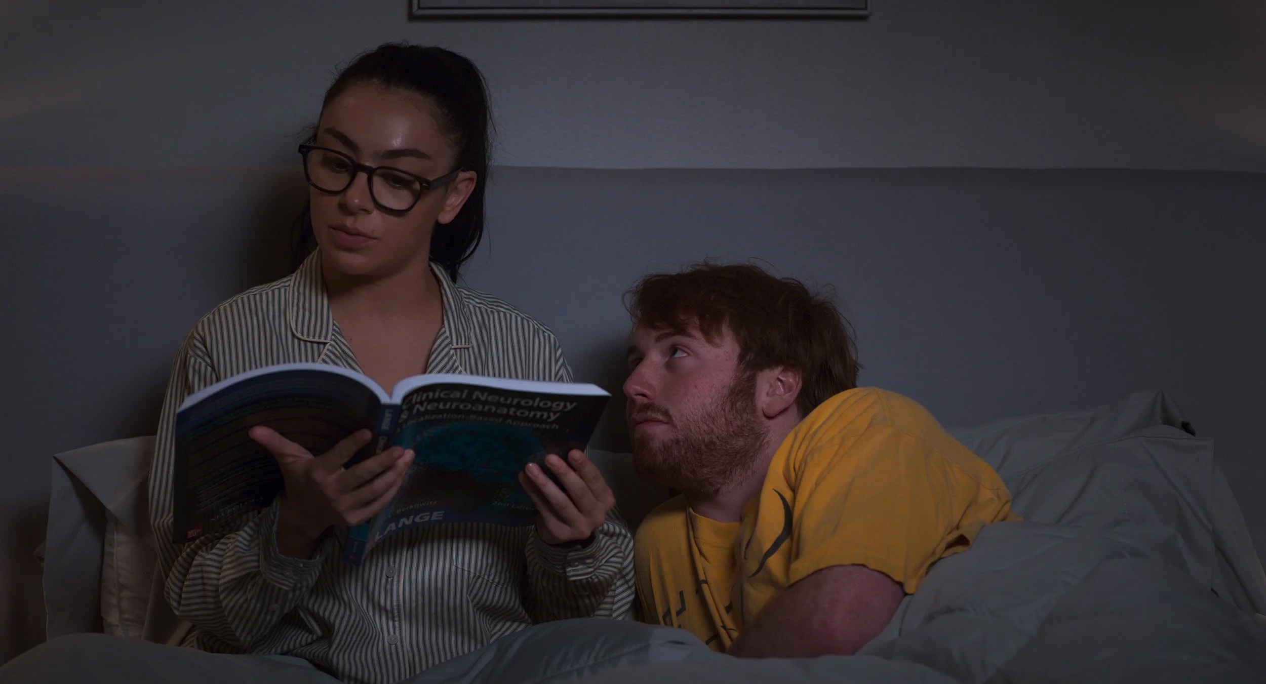 charli xcx as minerva reading a book in bed and cooper hoffman as elliot looking up at her in the movie i want your sex