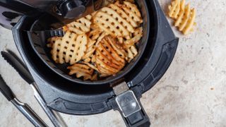 An air fryer basket with potato waffles in it next to silicone tongs