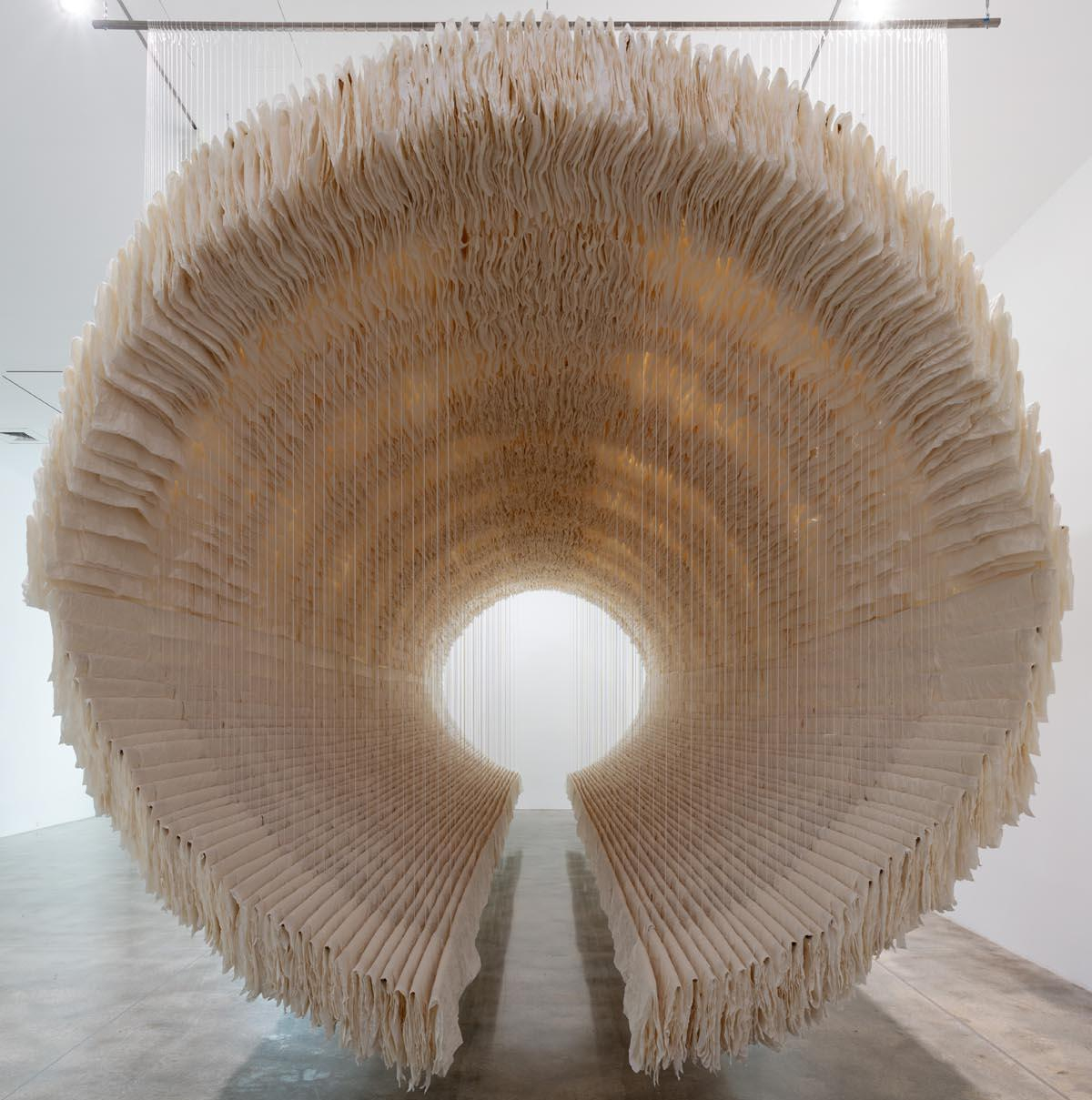 A round sculpture made of beige paper hangs in a brightly lit gallery.