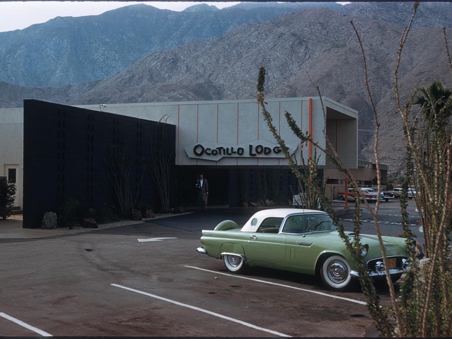 An old Californian mid-century modern hotel's facade, sat against a bare mountain range, features a vintage sign that reads "OCOTILLO LODGE", in front of which is parked a retro, green and white car.