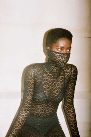 Model wears sheer black body suit, partly covering face