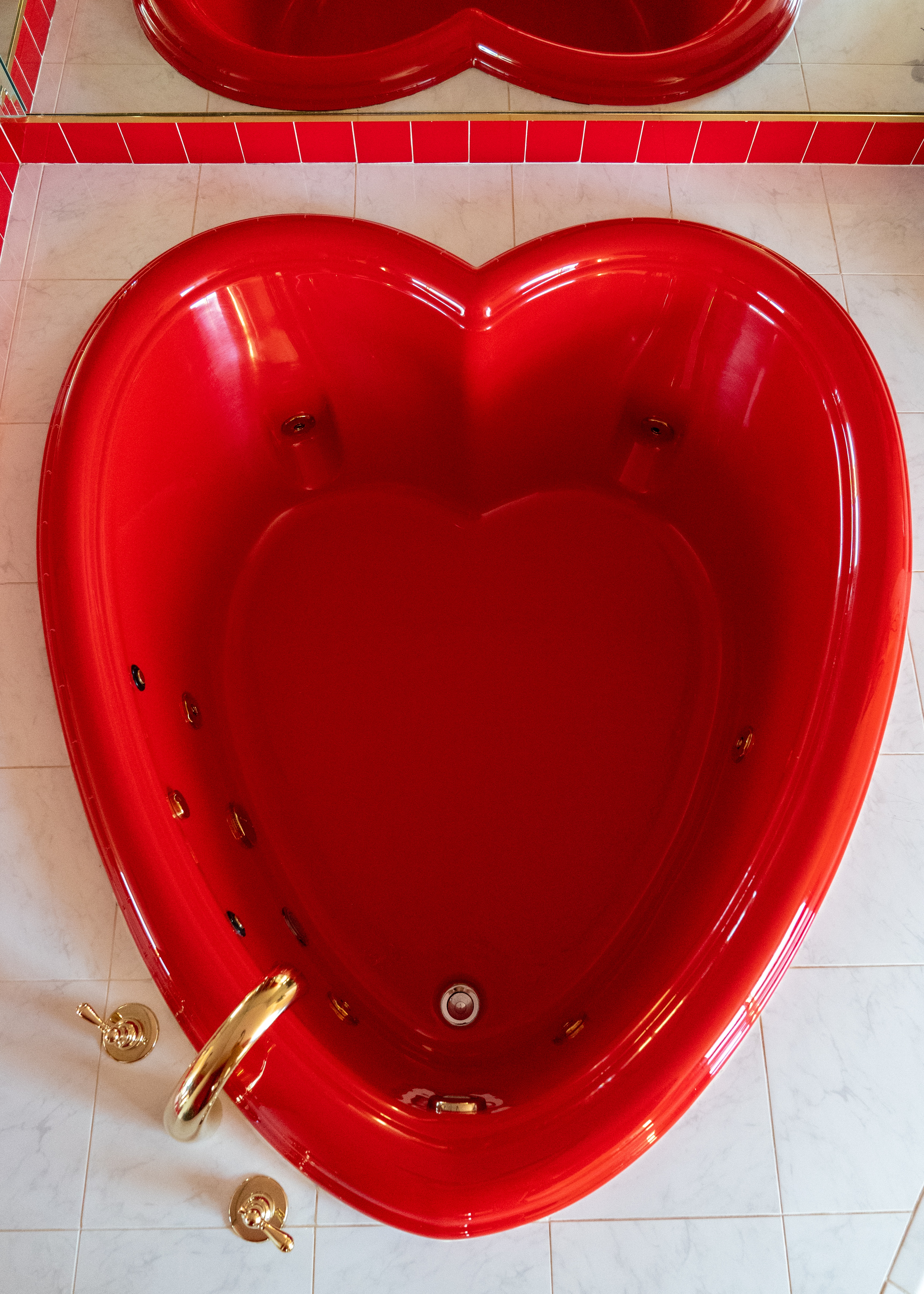 A heart-shaped bathrub in red and gold sits atop a series of white tiles in a bathroom.