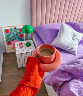A cup of tea in a coral cup, with a colorful bed with a pink headboard in the background