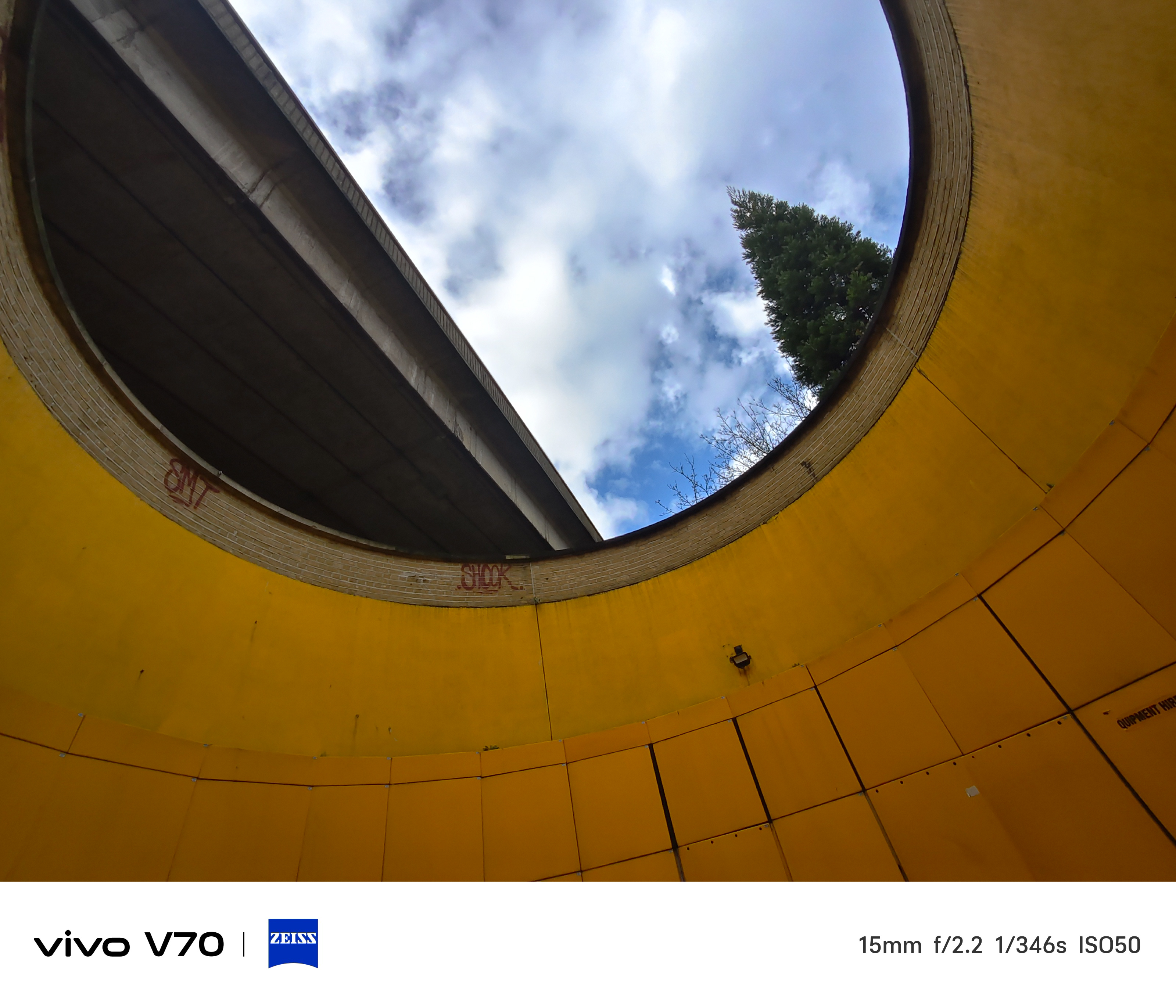 Ultra-wide shot looking up through circular yellow structure framing sky and overpass.