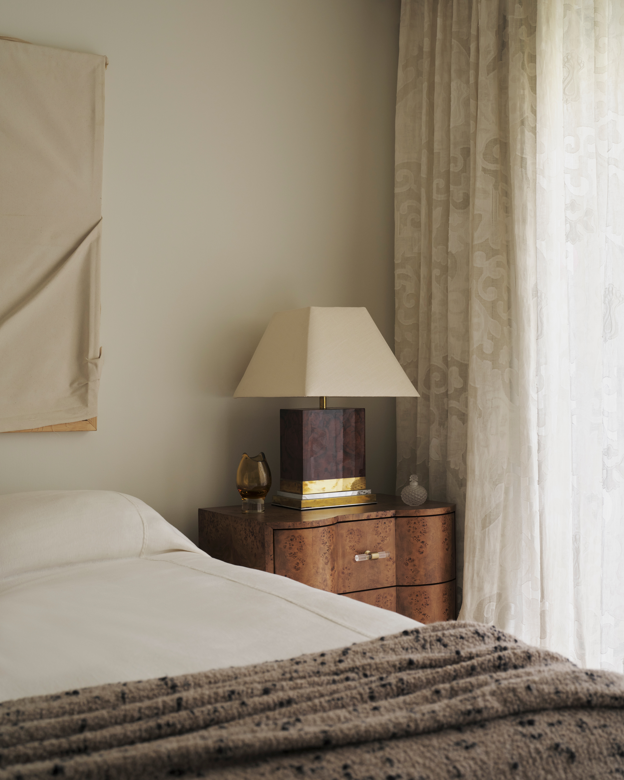 Image of a burl wood nightstand with a burl wood and gold lamp in a white bedroom. There are sheer, patterned curtains and a white bedspread and a textured beige throw.
