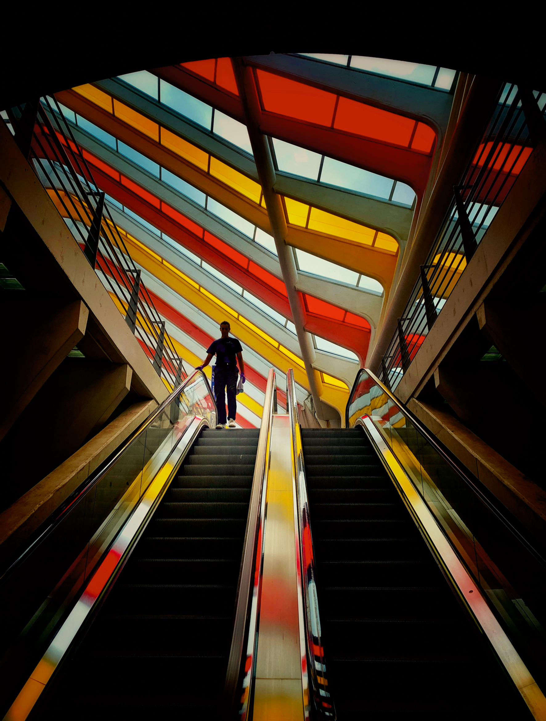 Looking up a pair of escalators towards a ceiling of diagonal red, orange, yellow and blue glass panels, a silhouetted man descends on the left-hand escalator.