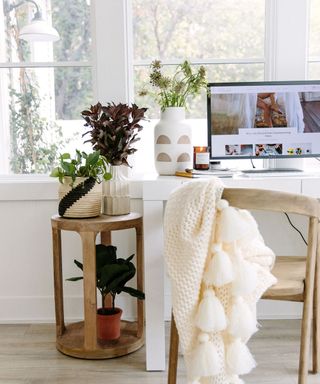 WFH set-up by window, with greenery in vases, natural materials, and chunky tasseled throw casually draped on wood chair.