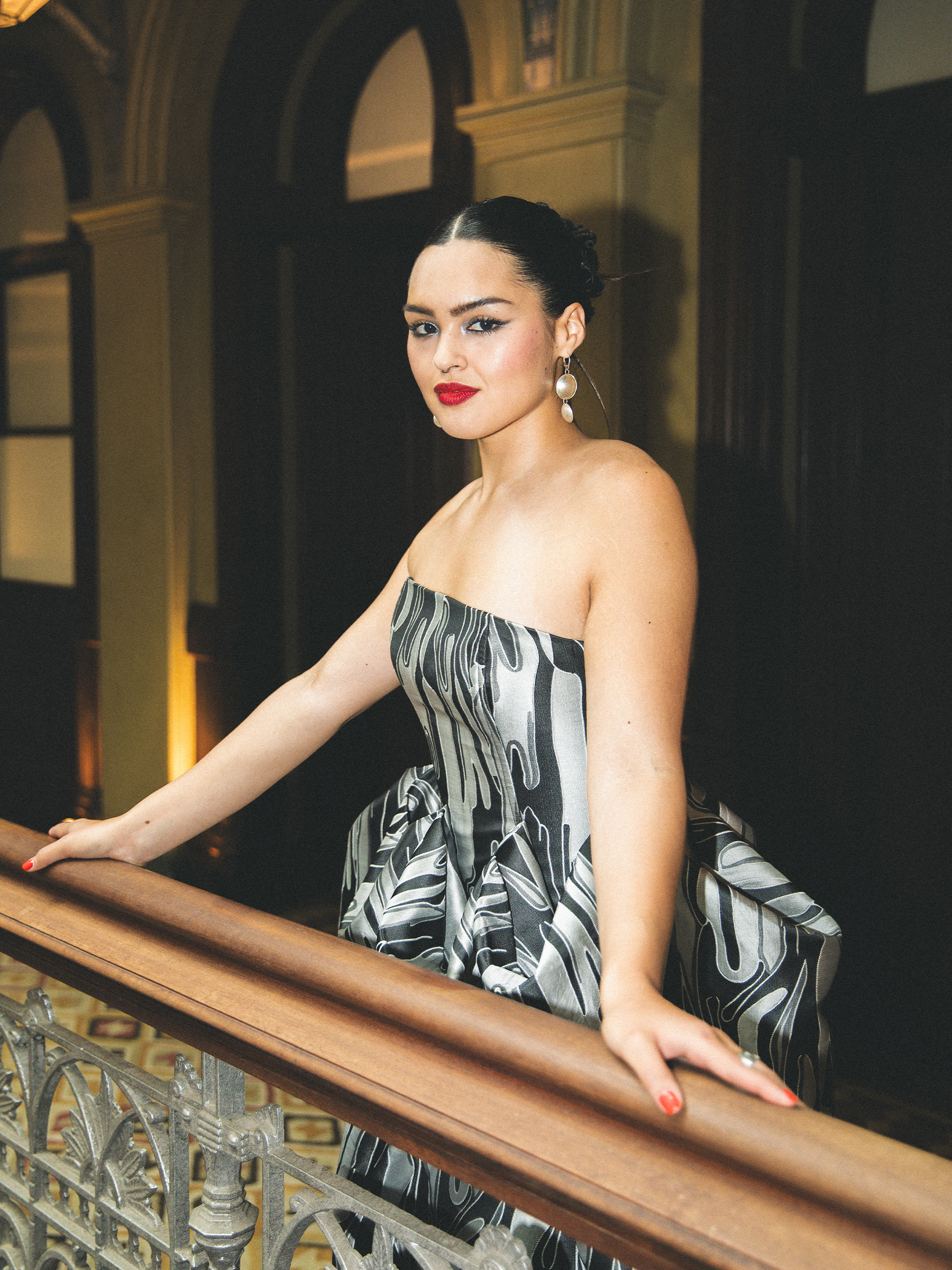 Anna Lambe stands against the railing of her hotel lobby. She is wearing a silver and black strapless gown with brocade design. Her hair is slicked back into a bun, and she has a red lip.