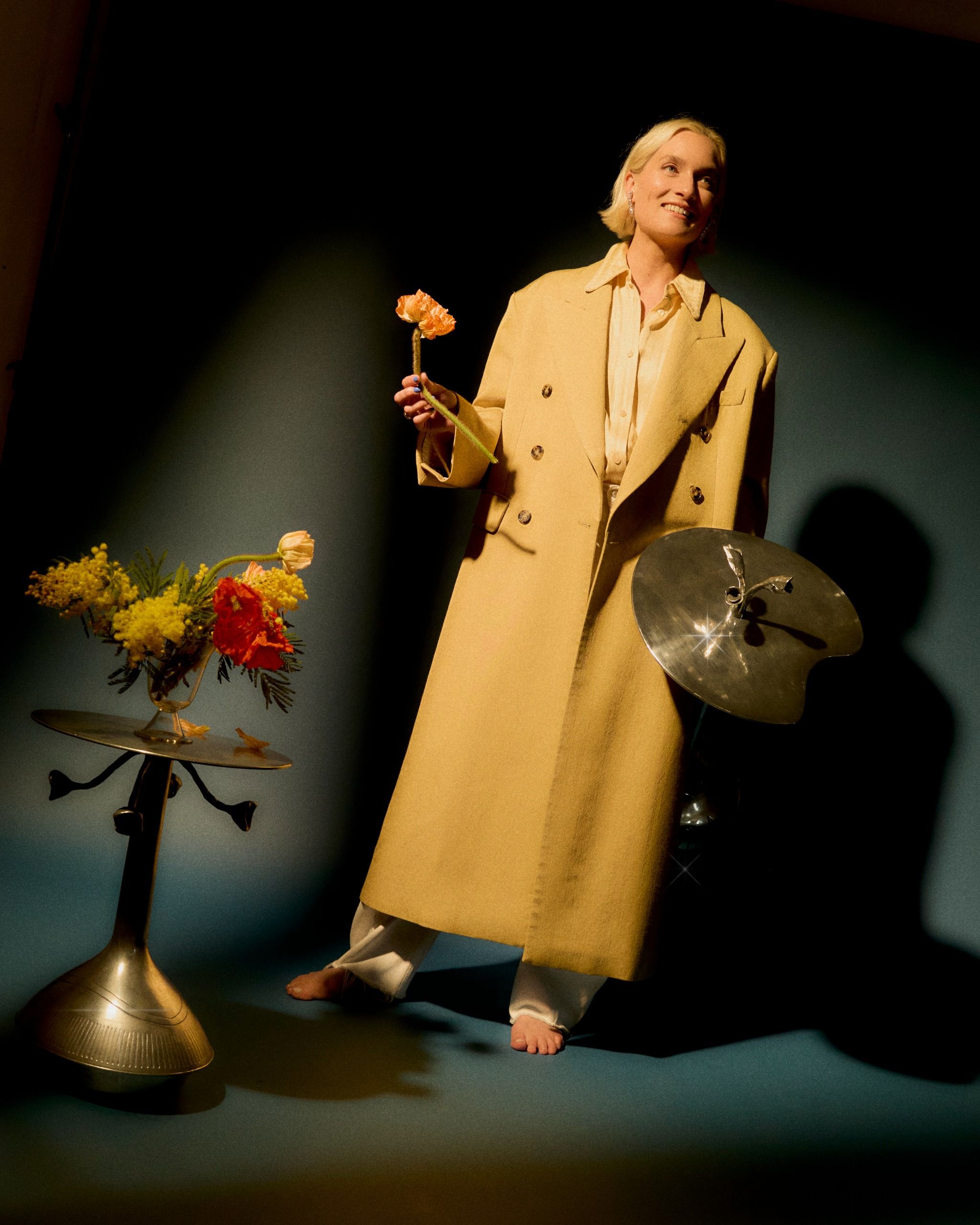 A young woman with blonde short hair, a long beige trench coat, white trousers, stands smiling barefoot in a blue set, surrounded by flowers styled in metallic vase.