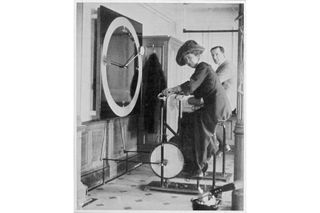 Best Exercise bikes were even found on the Titanic! This is an image from the boat with a woman sitting on an exercise bike, in a dress with a large hat looking at the camera. The front of the bike is facing to the left of the image, in front of her is a large clock which hands will move when she pedals. There is a man in a suit behind her also sitting on a bike.