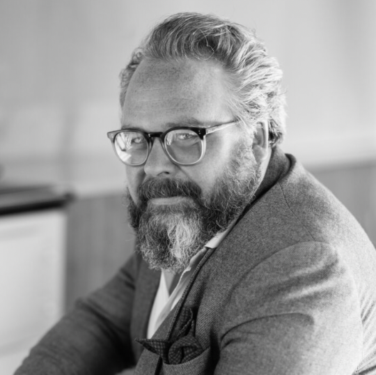 A grayscale headshot of a man with glasses and a beard