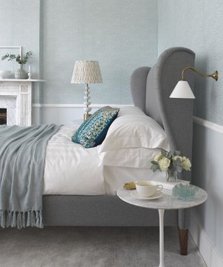 An example of how to plan bedroom lighting showing a grey bed with a white bedside lamp and a white and gold wall lamp