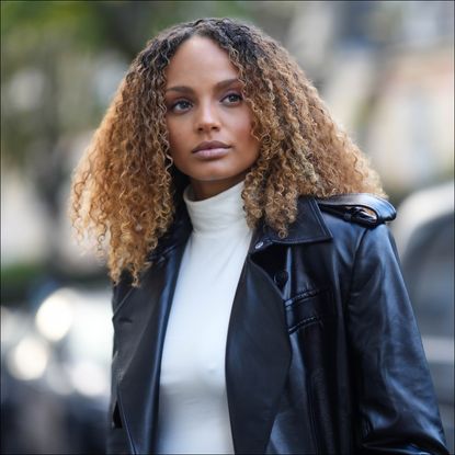 Alicia Aylies has curly hair and wears a white sleeveless / high neck body t-shirt, a black shiny leather varnished long trench coat, during a street style fashion photo session, on November 09, 2022 in Paris, France