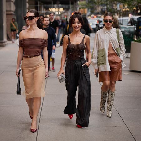 Women are walking and posing in stylish outfits. One wears a skirt and off the shoulder top. The next wears a tank top and trousers. The last wears leather shorts and button down shirt with boots.