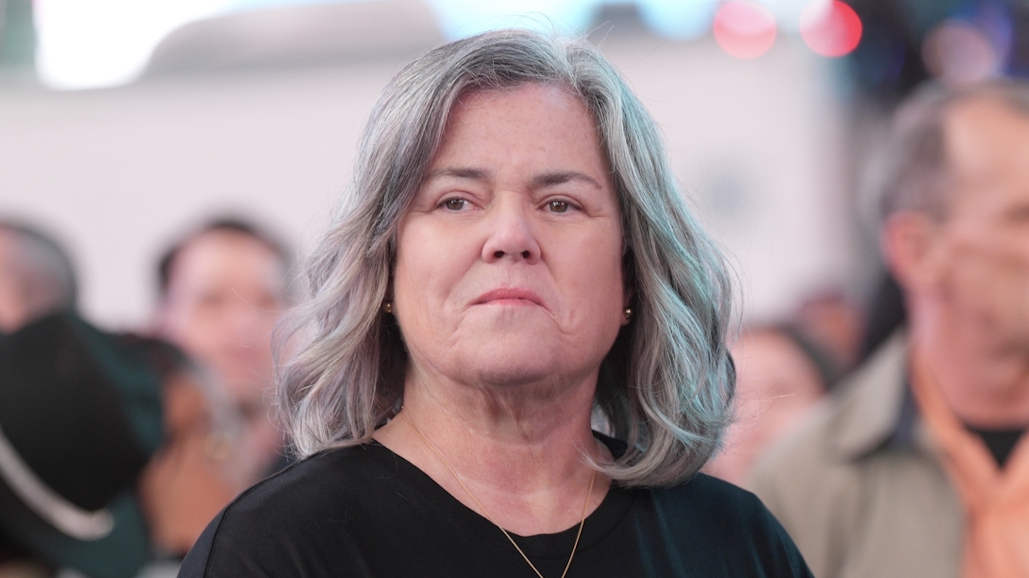 Rosie O'Donnell with shoulder-length gray hair standing in a crowd in Hacks Season 4