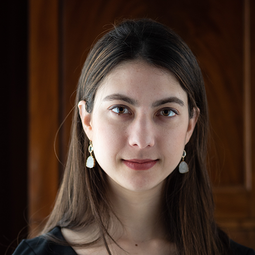 A woman with long dark hair and brown eyes, wearing dangling earrings looks at the camera