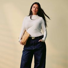 model wears long-sleeve white blouse, dark wash baggy jeans, and a tan clutch while posing for a picture and hair blows in the wind. 