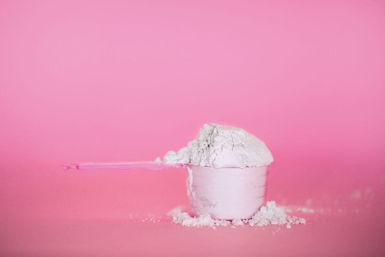 A pink spoon measuring protein powder against a pink background. 