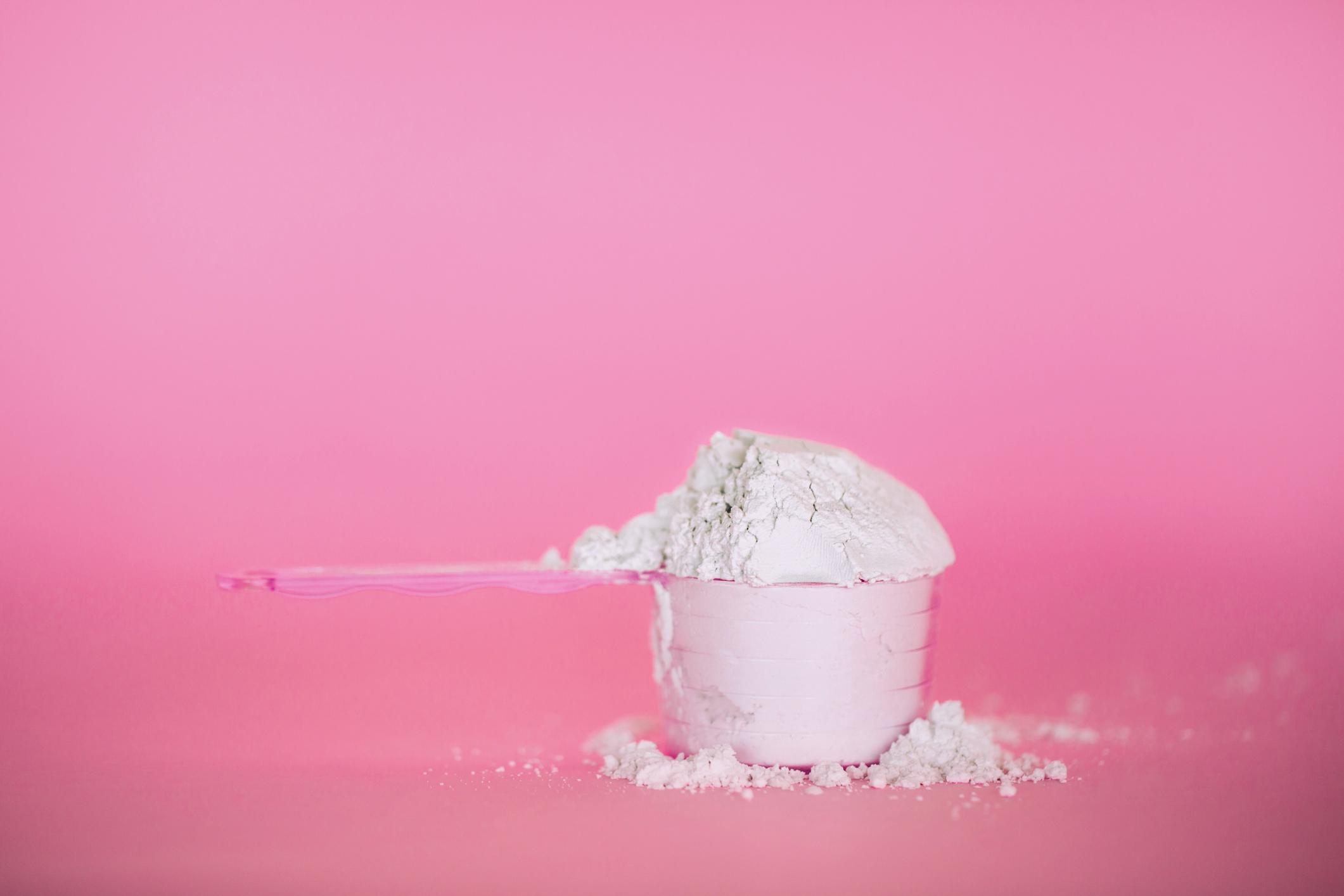  A pink spoon measuring protein powder against a pink background.  