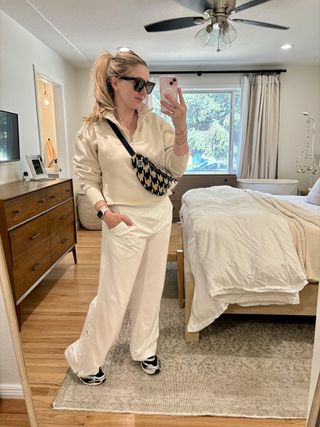 woman in bedroom wearing white pullover zip-top sweatshirt, white sport pants, black-and-white trainer sneakers, black-and-white houndstooth fanny pack, black sunglasses