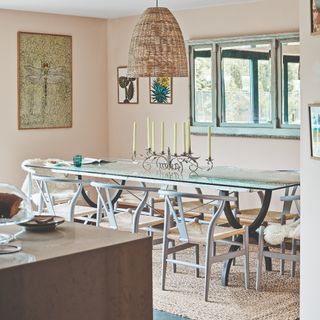 A soft peach-painted dining room with a long rectangular glass dining table and an oversized rattan pendant light above it