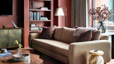 living room with deep red walls, beige sofa with cushions, brass console with sculpture on top, timber coffee table, lamp and bookshelf