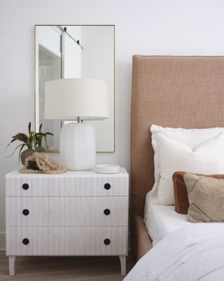 Night stand with table lamp and mirror above next to bed with upholstered headboard