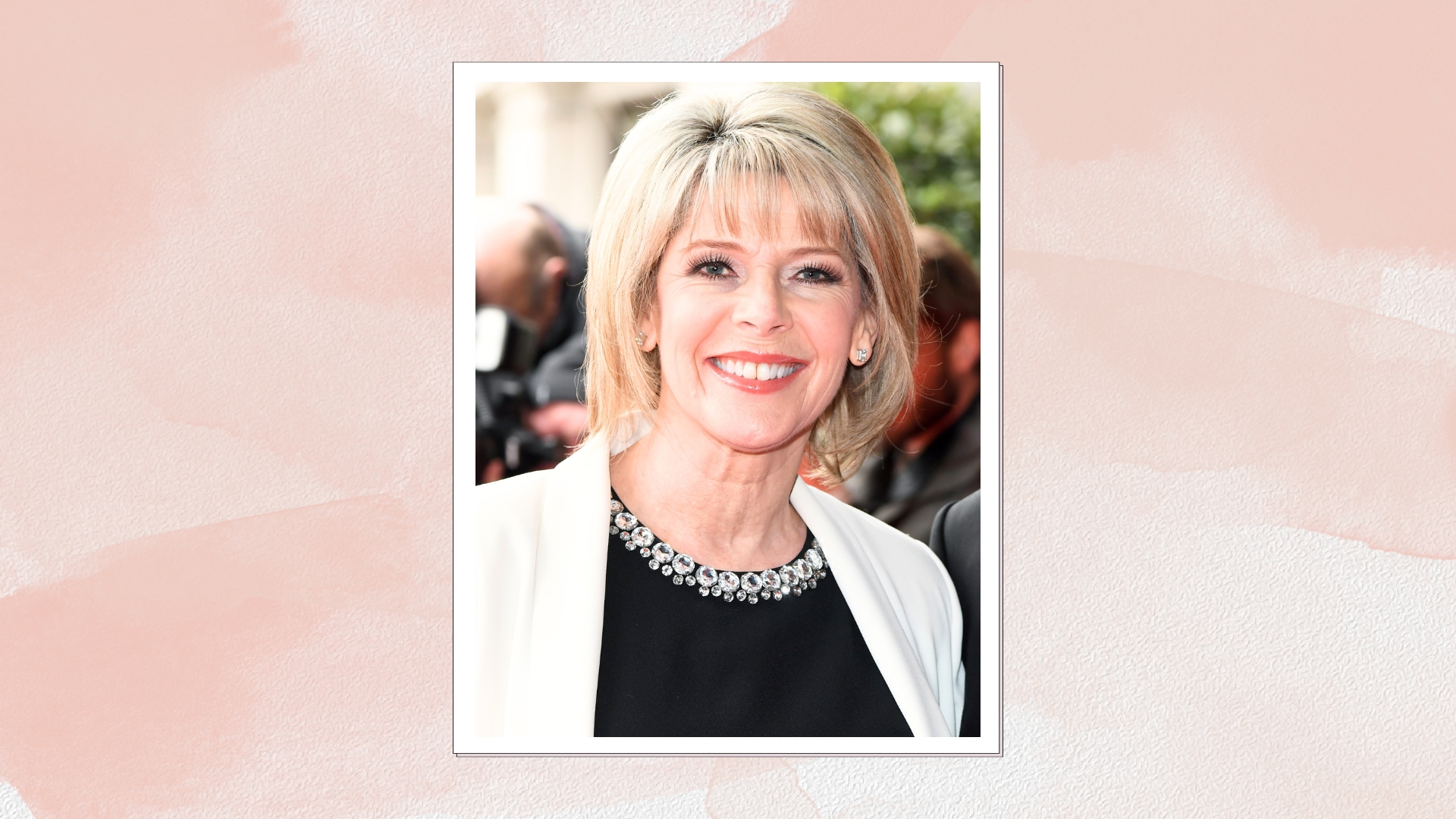 Ruth Langsford smiling with blonde bob and fringe, wearing a black crystal-embellished neck top and white blazer, on a pink watercolour background