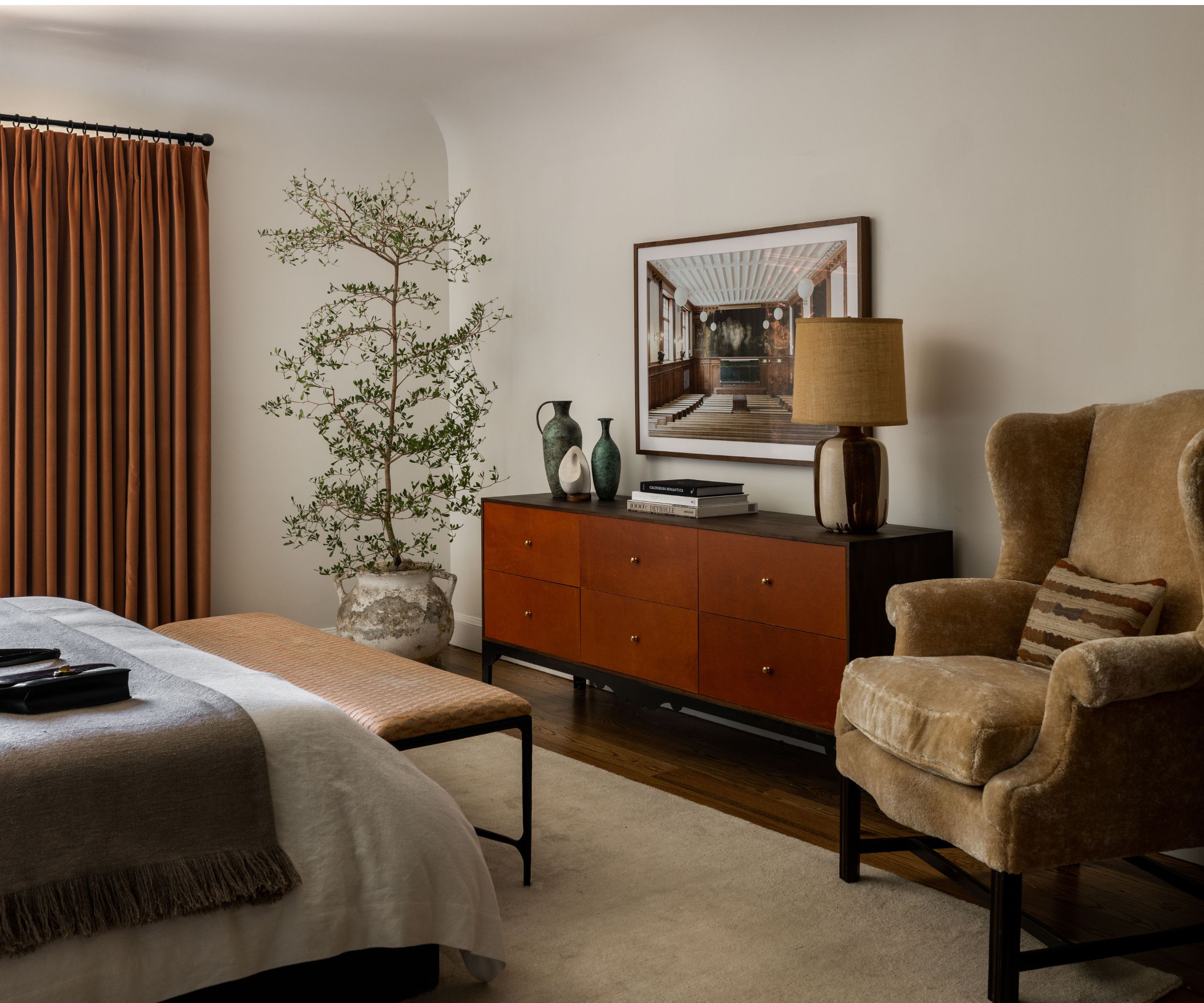 neutral cozy modern bedroom with wooden floor, a large deep pile rug, burnt orange curtains, a neutral styled bed with a black and teak woode sideboard, plant, and sheepskin chair