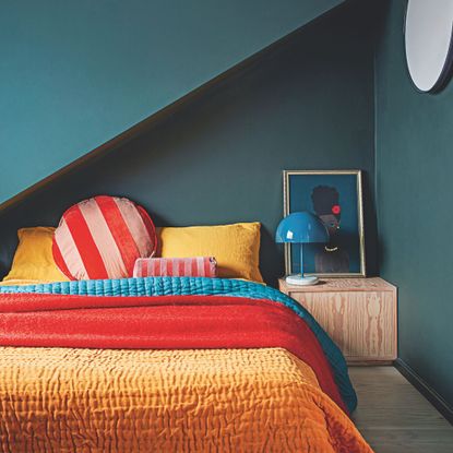 Blue painted bedroom with orange and red bedding on the bed, and a wooden side table