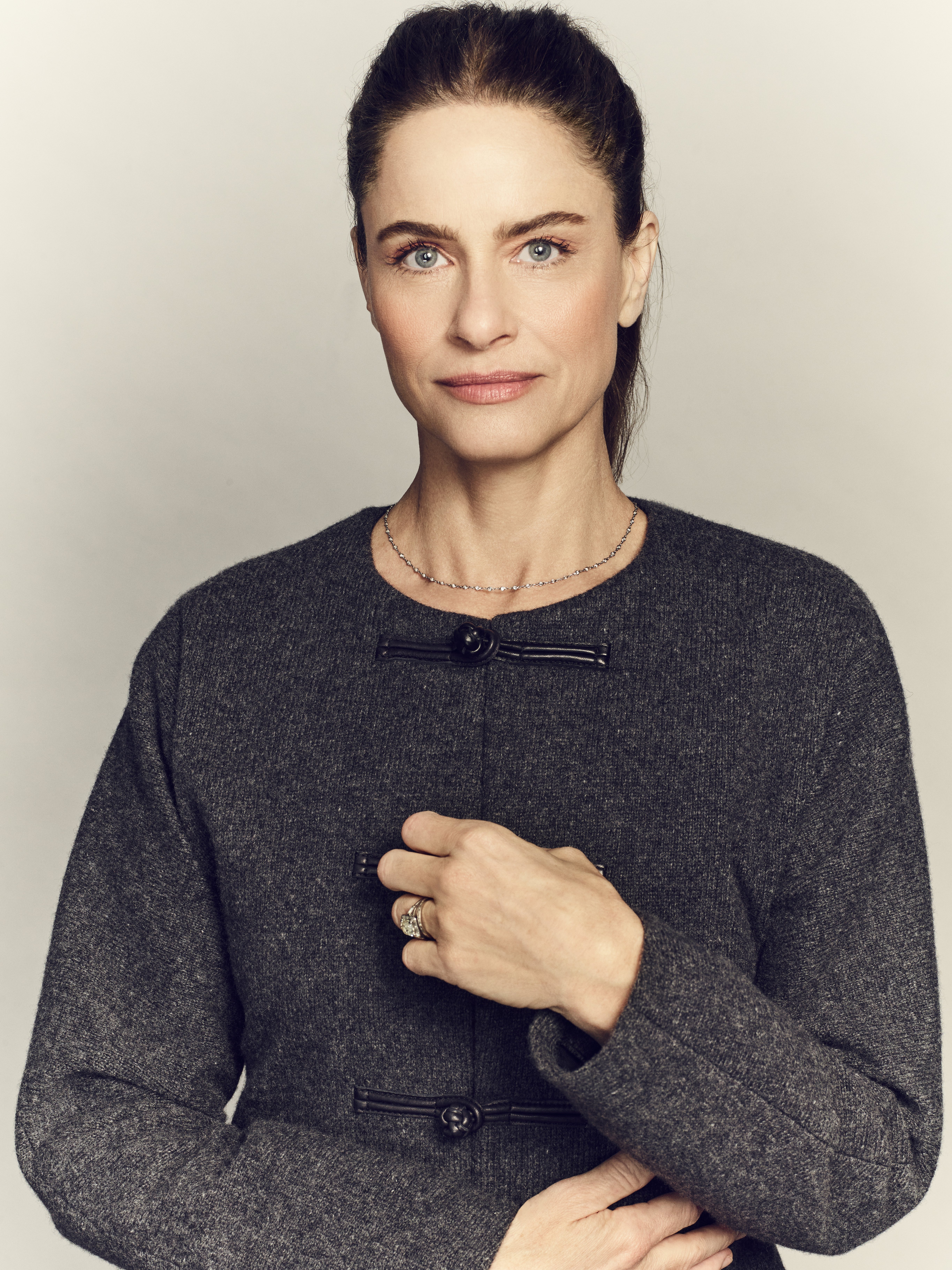 amanda peet poses wearing a grey jacket and her hair in a ponytail in a promotional shot for your friends and neighbors season 2