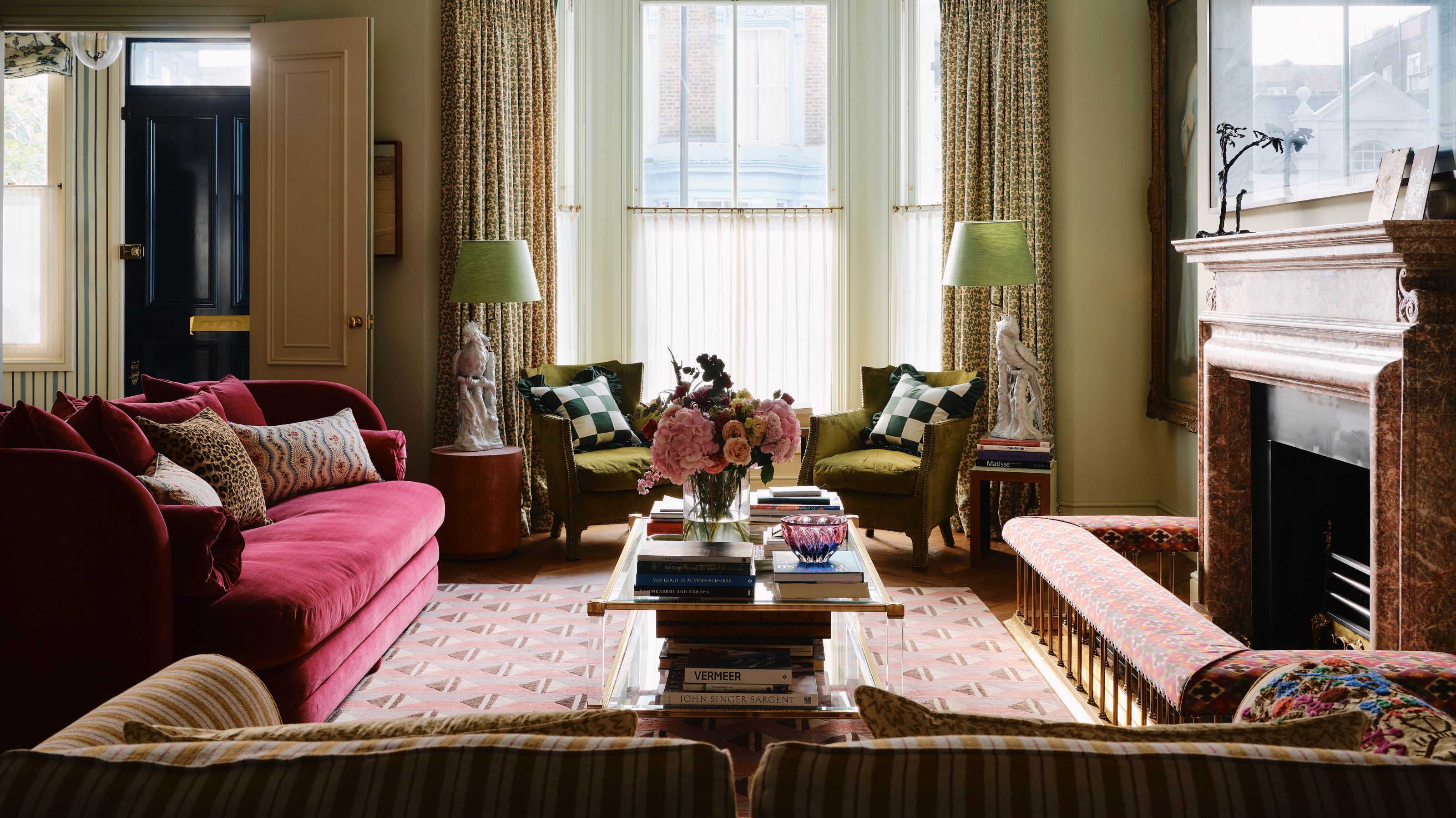 warm neutral english traditional living room with a large bay window, fireplace with a fender, dark red velvet couch, two green armchairs with side tables and lamps