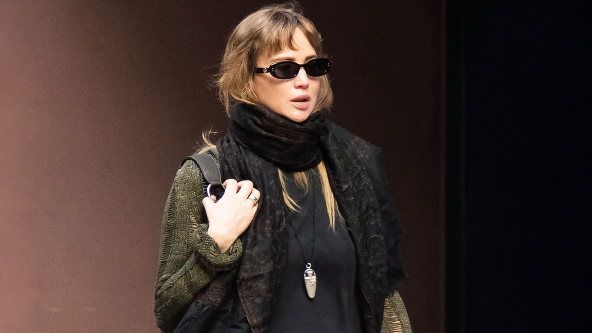 Jennifer Lawrence walks down the street in New York wearing black split-hem trousers with rust red satin shoes a black top, an oversized scarf and a green cardigan.