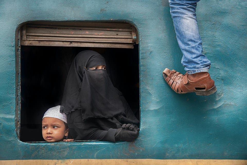 A woman wearing a burqa looking out of a train window with a child. A man&#039;s leg and foot is visible as he climbs on to the roof 