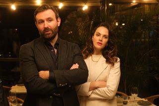 Miles (James McArdle) and Lucy (Jessica Brown Findlay) stand in a restaurant with fairy lights suspended across the ceiling. They are both wearing expensive-looking suits, Miles in black and Lucy in white, and both have their arms folded and look unimpressed.