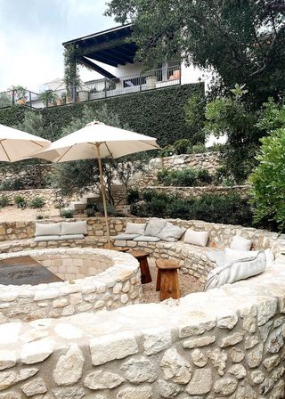 Circular stone sloped backyard ideas with integrated seating and firepit, below a high terrace with climbing plants and olive trees.