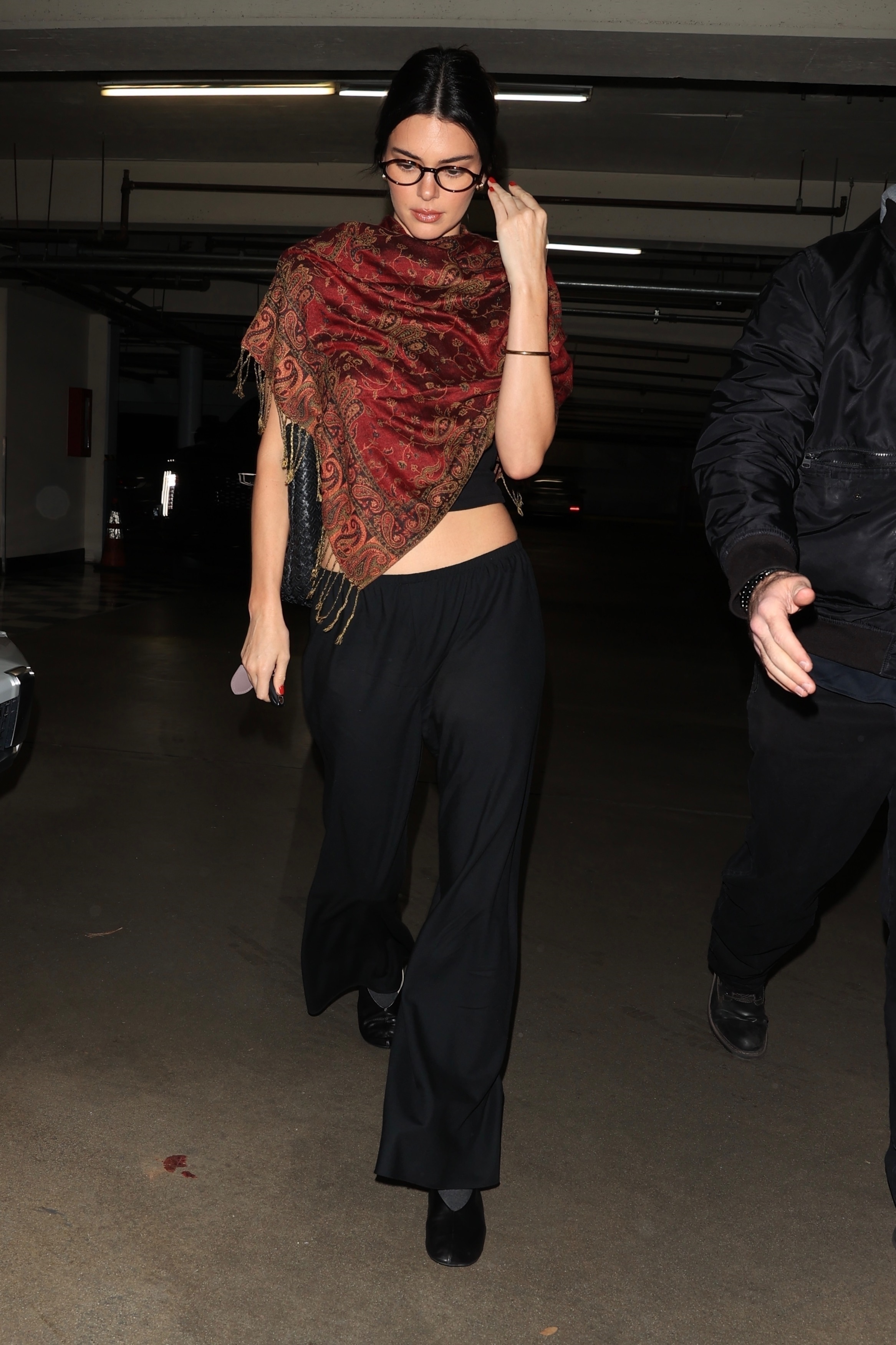 Kendall Jenner wears a printed scarf over a black tank top styled with black pull-on pants and black ballet flats.