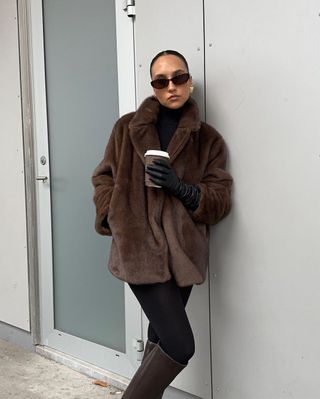 A woman wears a brown fur coat, brown sunglasses, black leggings, and brown-knee high boots.