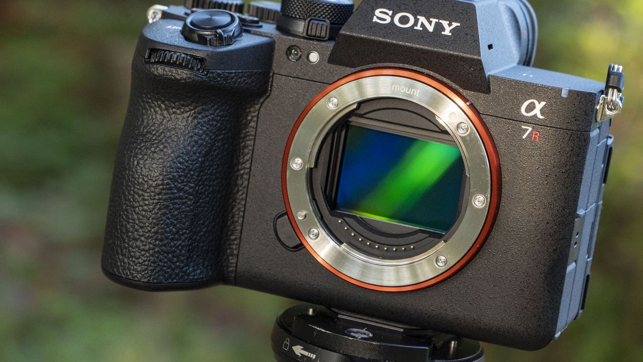 The Sony A7R VI camera and no lens attached and the sensor inside is reflecting light, it's mounted to a tripod outdoors, there's a natural background