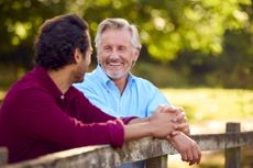 Mature Father With Mixed Race Adult Son Leaning On Fence Walking In Countryside.