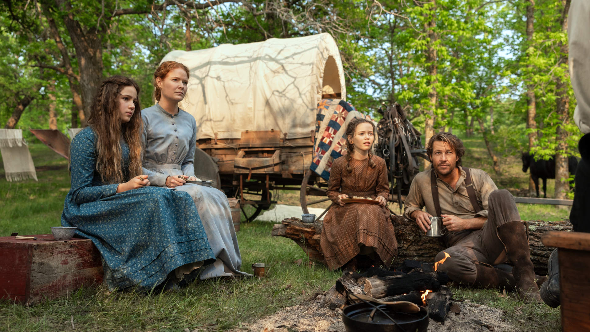 The Ingalls family (Skywalker Hughes as Mary, Crosby Fitzgerald as Caroline, Alice Halsey as Laura, Luke Bracey as Charles) sits around a campsite with a wagon behind them, in 'Little House on the Prairie.'