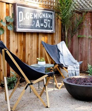 A backyard with a wooden fence with a black and white chalkboard on it, and four chairs around a black fire pit