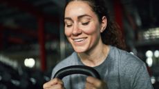 The camera frames smiling woman holding the horns of a kettlebell 