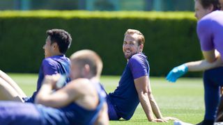 Harry Kane smiling at the camera while sitting on the pitch with teammates.