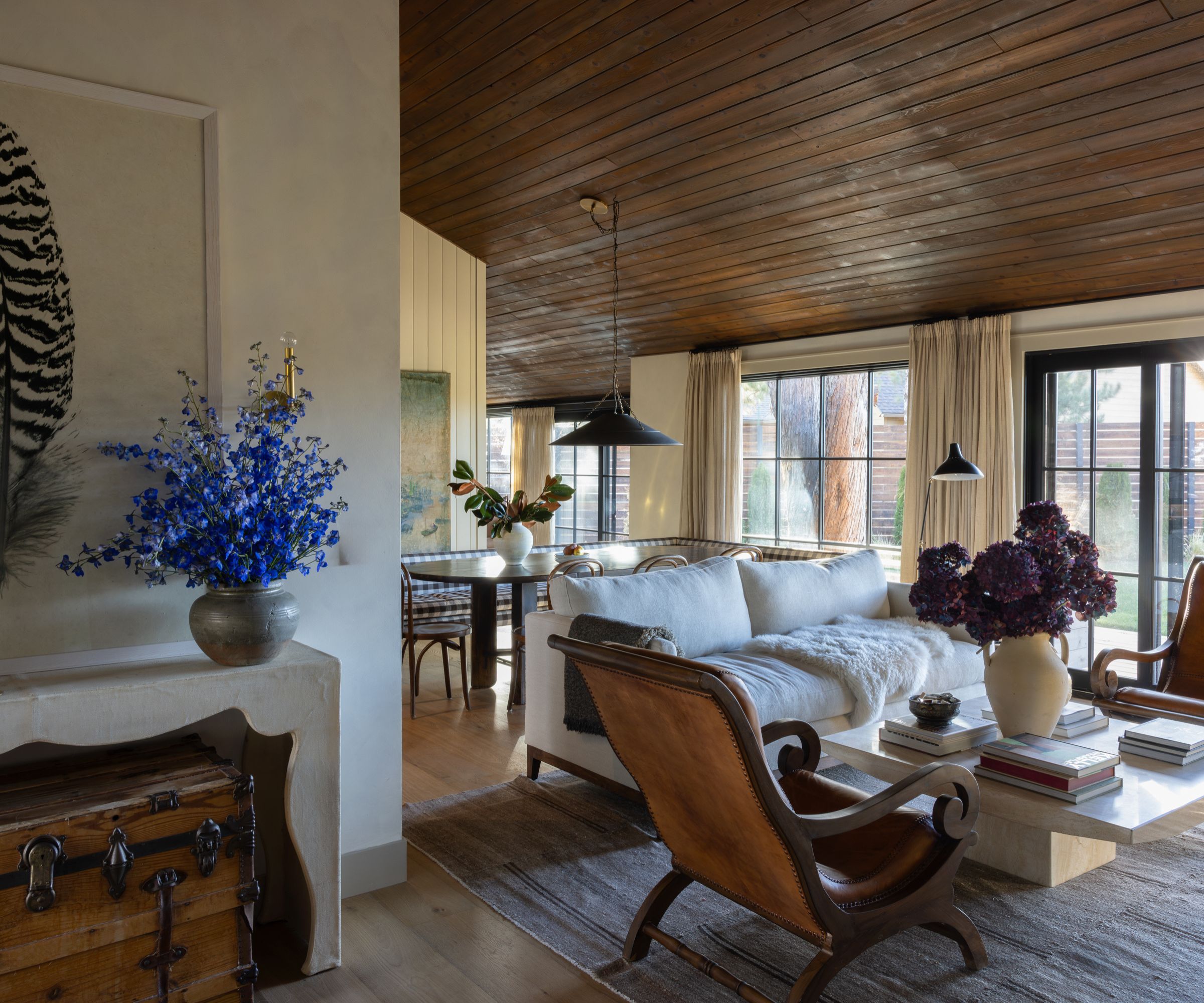 View into a cozy living area with a white sofa, leather chairs, and a coffee table with books and purple flowers. The dining area with a checkered banquette and large windows can be seen in the background.