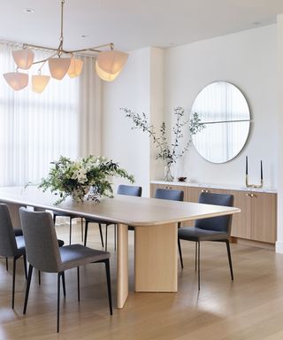 Dining room with light wood table, gray upholstered chairs and sideboard, pendant light, mirror, and foliage and flower arrangements