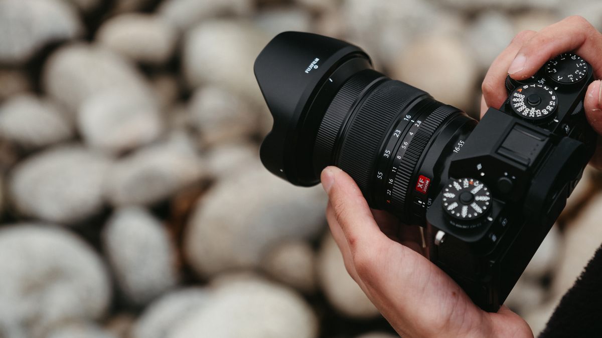 Fujifilm XF 16-55mm f/2.8 R LM WR II lens attached to a camera and held in a pair of hands outside over large pebbles