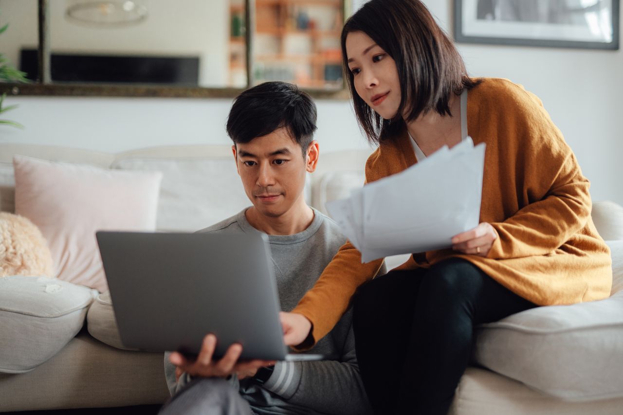 A couple sits on a couch with papers and a laptop reviewing their finances.