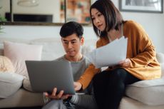 A couple sits on a couch with papers and a laptop reviewing their finances.